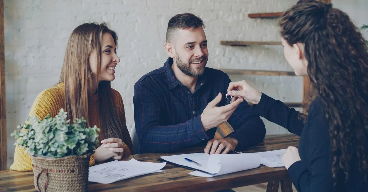 Un couple souriant reçoit les clés de sa maison des mains d'une agente immobilière assise à un bureau en bois.