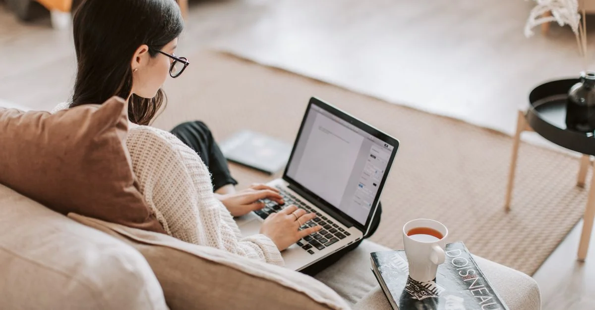 Femme travaillant à distance sur un ordinateur portable depuis un canapé confortable dans un salon, savourant une tasse de thé.