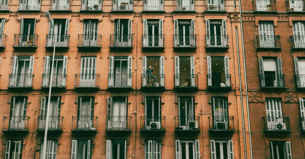 Un immeuble d'appartements en briques avec plusieurs étages, avec des balcons et un résident visible sur l'un d'eux.