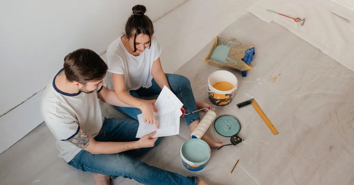 Un couple assis sur le sol, examinant des documents au milieu de matériaux de rénovation domiciliaire.