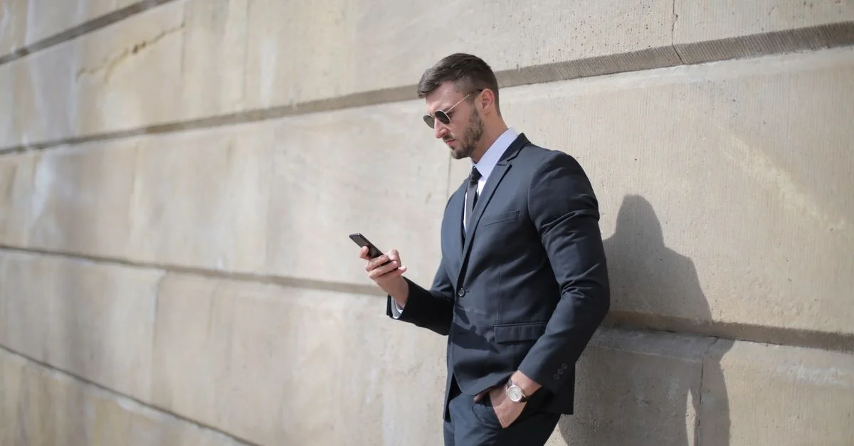 Homme confiant en costume noir s'appuie sur un mur tout en utilisant un smartphone à l'extérieur.