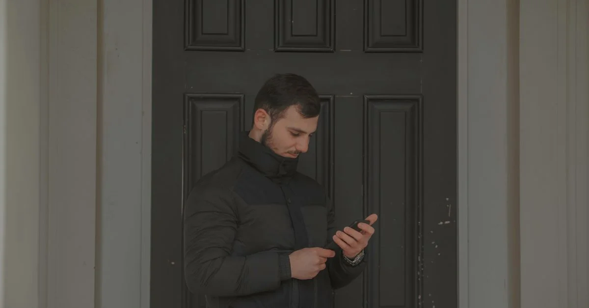 Un homme en veste noire se tient près d'une porte sombre, absorbé par son smartphone.