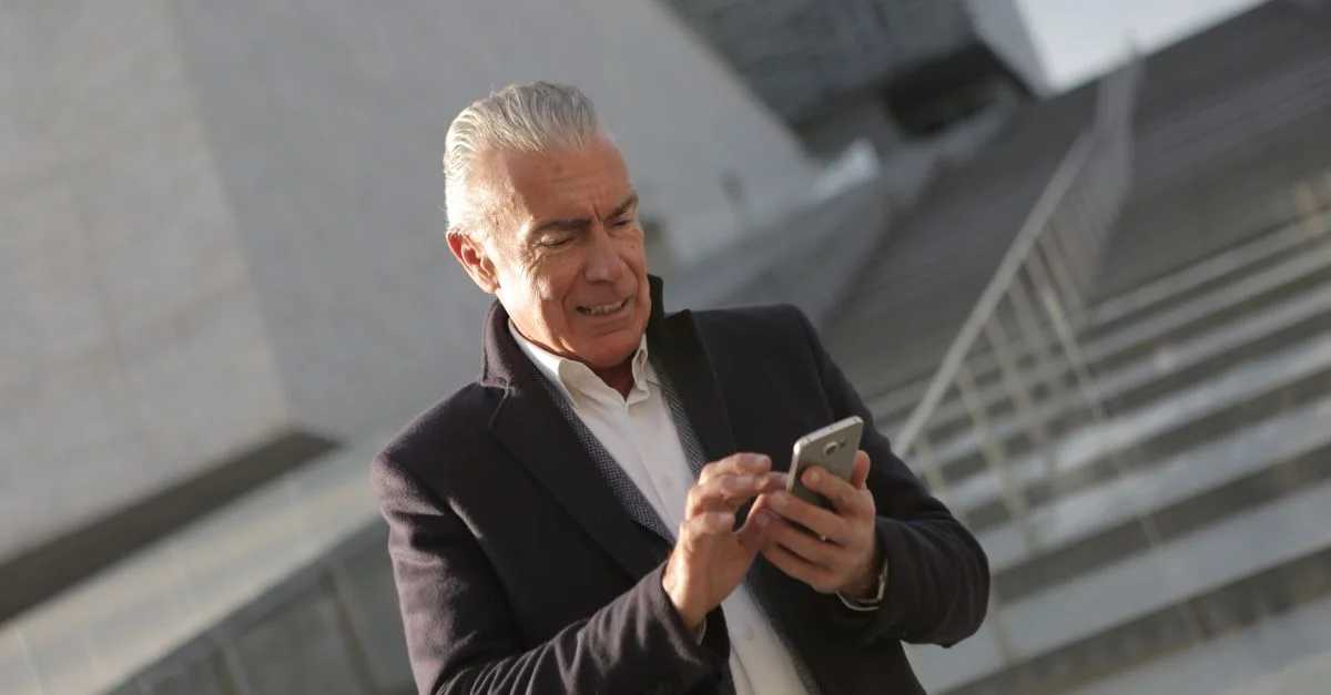 Homme âgé en tenue professionnelle utilisant un smartphone en extérieur devant un bâtiment moderne.
