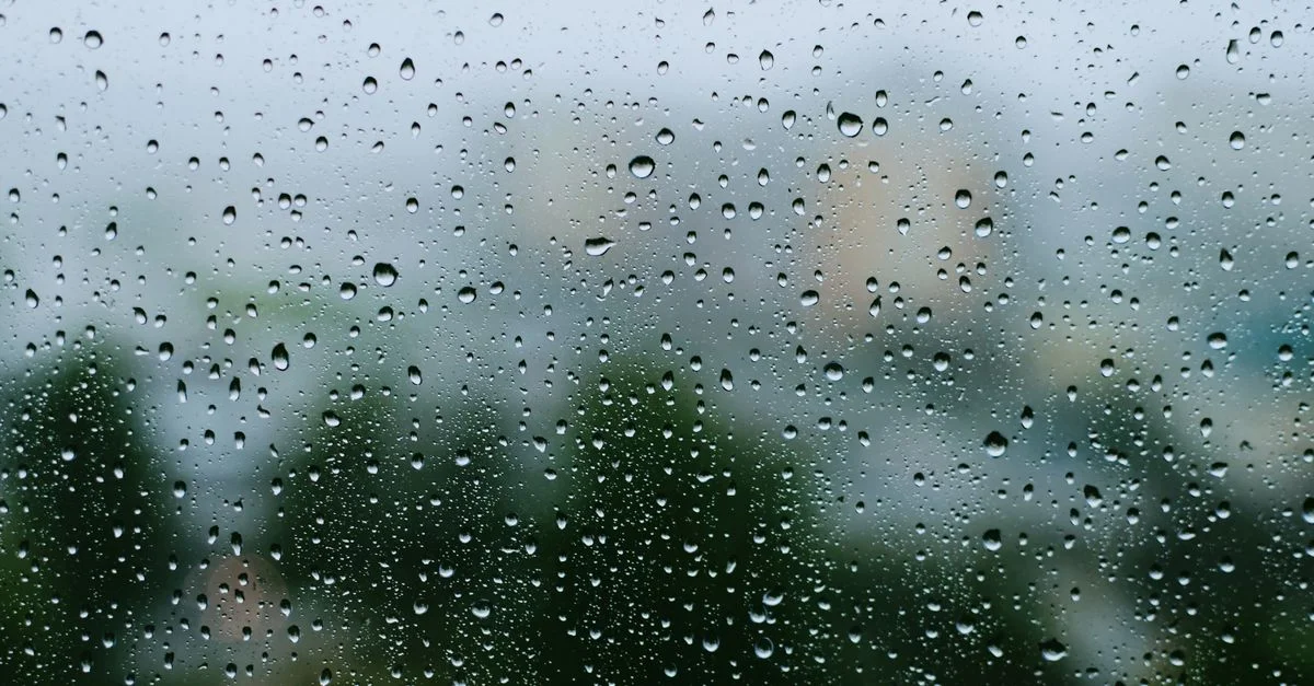 Vue en gros plan des gouttes de pluie sur une fenêtre avec un paysage urbain flou en arrière-plan, capturant l'ambiance d'une journée pluvieuse.