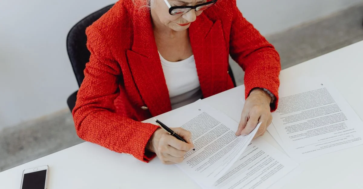 Femme d'affaires âgée en veste rouge signant des documents à son bureau, montrant son leadership.